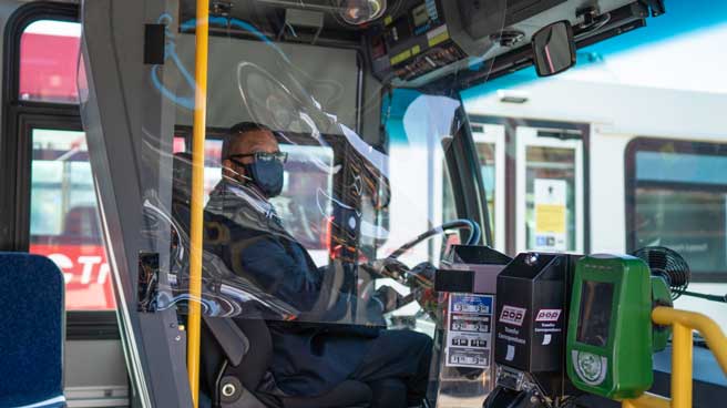 OC Transpo Operator seated at the wheel of a bus wearing a cloth face mask. The Operator area is protected by a soft shield made of clear plastic.