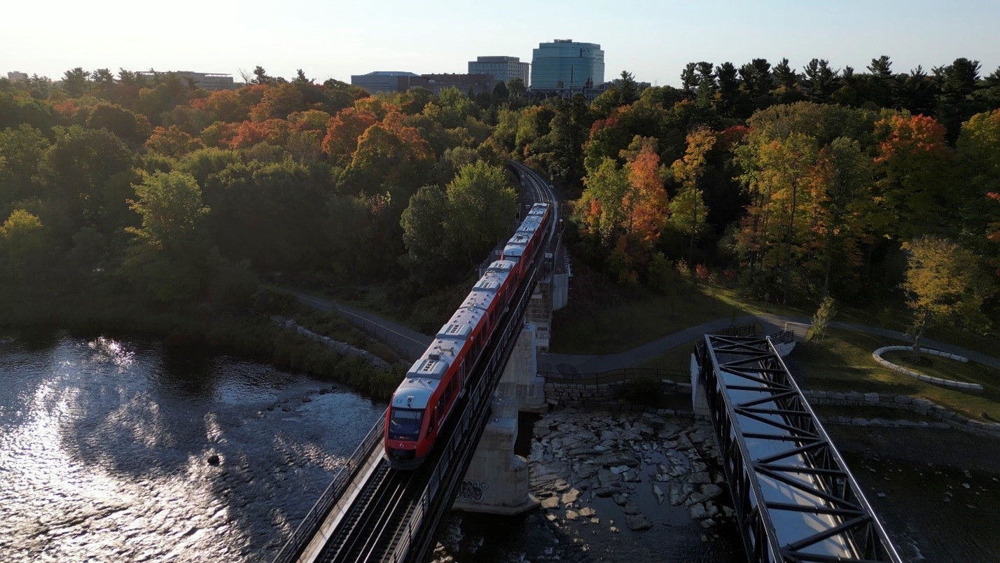 Un train Stadler FLIRT traversant le pont près de l’Université Carleton au-dessus de la rivière Rideau, avec des arbres d’automne et des bâtiments de la ville en arrière-plan.